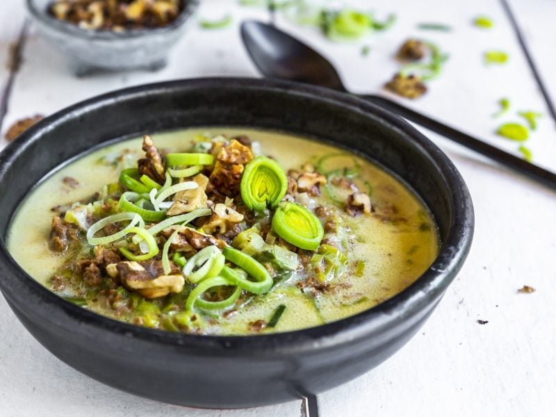 Draufsicht, schräg: Eine schwarze Schüssel Käse-Lauch-Suppe mit Walnüssen auf einem weißen Untergrund aus Holzdielen. Daneben liegen ein Suppenlöffel, lauchringe und eine Schale mit Walnusskernen.