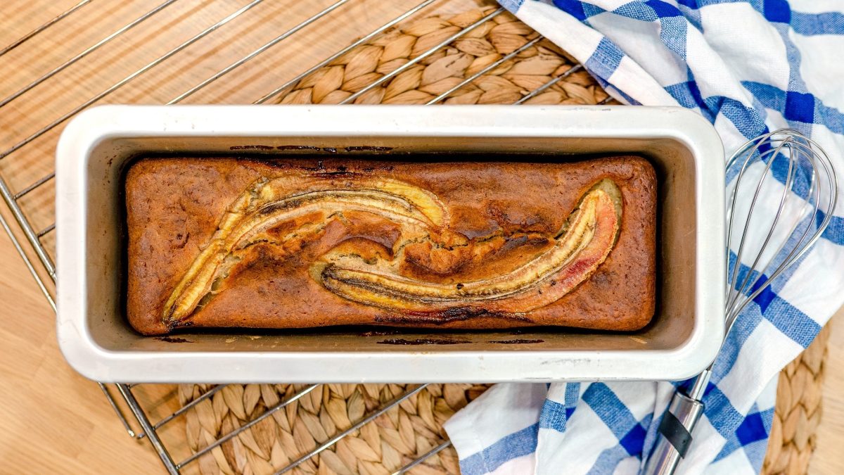 Draufsicht: Ein Low Carb-Bananenbrot in einer weißen Backform auf einem hellen Unterrund aus Holz. Daneben liegt ein Küchenhandtuch.