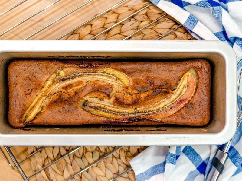 Draufsicht: Ein Low Carb-Bananenbrot in einer weißen Backform auf einem hellen Unterrund aus Holz. Daneben liegt ein Küchenhandtuch.