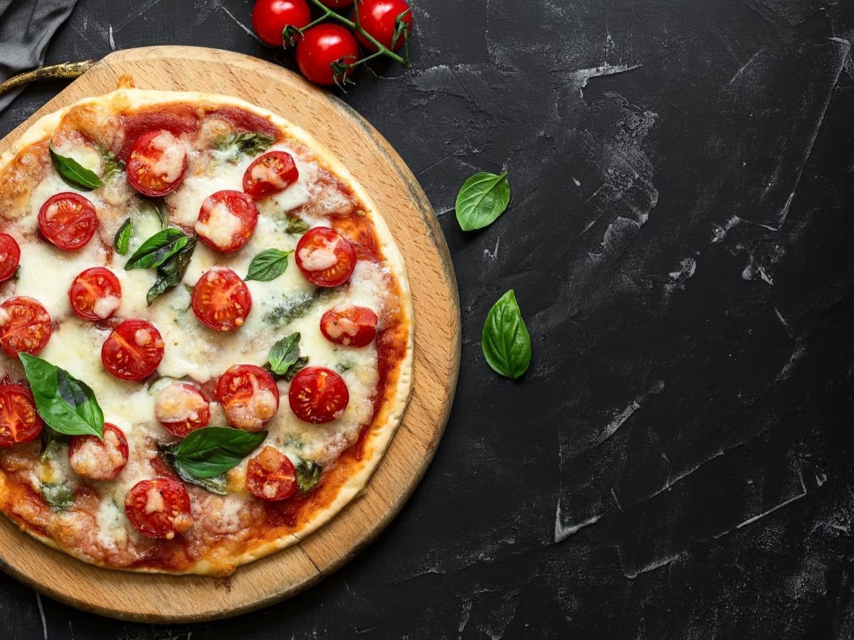 Draufsicht: Auf dunklem Untergrund liege eine Low-Carb-Pizza ohne Mehl. Die Pizza Margherita wurde mit einem Low-Carb-Pizzateig mit Quark gebacken. Sie ist mit Mozzarella belegt. Drum herum liegen frische Zutaten, wie etwa Basilikumblätter.