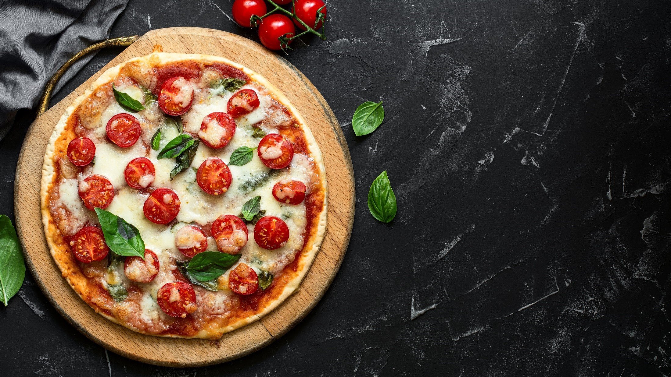 Draufsicht: Auf dunklem Untergrund liege eine Low-Carb-Pizza ohne Mehl. Die Pizza Margherita wurde mit einem Low-Carb-Pizzateig mit Quark gebacken. Sie ist mit Mozzarella belegt. Drum herum liegen frische Zutaten, wie etwa Basilikumblätter.