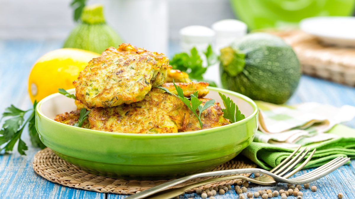 Frontal: Sommerliche Anmutung: Kohlenhydratarme Zucchinipuffer nach einem Low-Carb-Rezept liegen auf einem hellblauen Holztisch. Im Hintergrund sind frische Zucchini und andere Zutaten zu sehen.