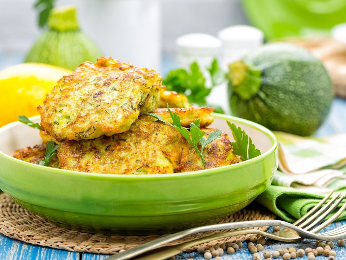 Frontal: Sommerliche Anmutung: Kohlenhydratarme Zucchinipuffer nach einem Low-Carb-Rezept liegen auf einem hellblauen Holztisch. Im Hintergrund sind frische Zucchini und andere Zutaten zu sehen.