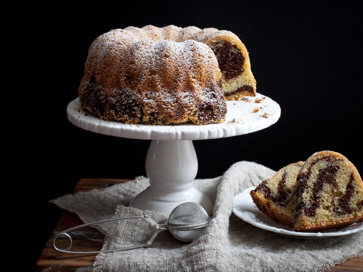 Frontal: Schwarzer Hintergrund. Eine Weiße hohe Prässentierplatte aus Porzellan, auf dem ein Marmor-Gugelhupf steht. Daneben ein weißer Teller mit einem Stück Kuchen.