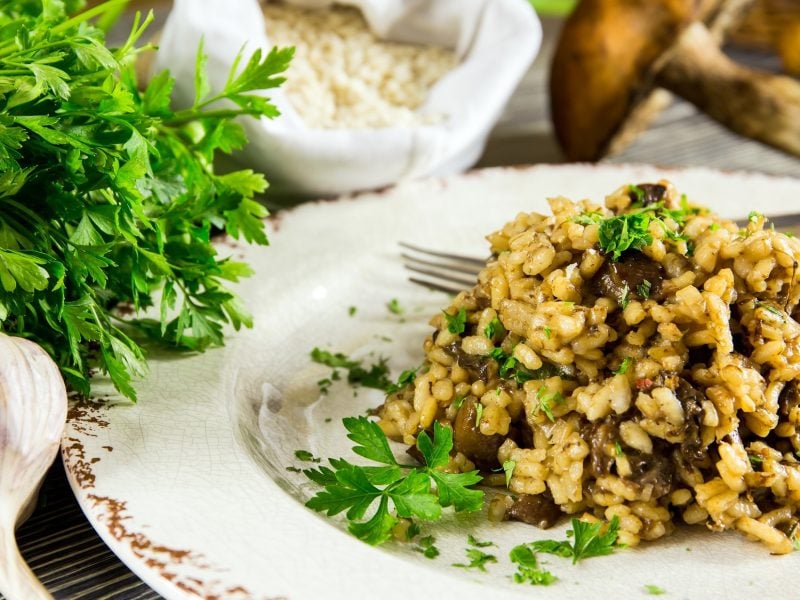 Frontal, leicht schräg von oben: Ein weißer Teller Risotto mit Waldpilzen. Daneben liegen eine Knoblauchknolle und ein Bund frische Petersilie. Im Hintergrund sind frische Pilze.