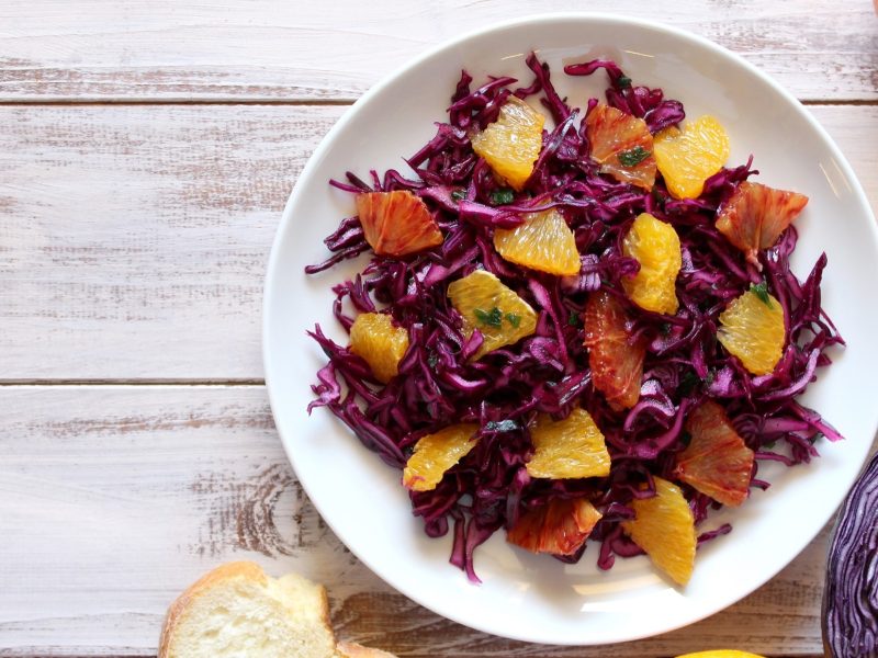 Draufsicht: Ein weißer Teller mit Rotkohlsalat mit Orangen auf einem weißen Untergrund aus Holz. drum herum liegen drei halbierte Zitrusfrüchte.