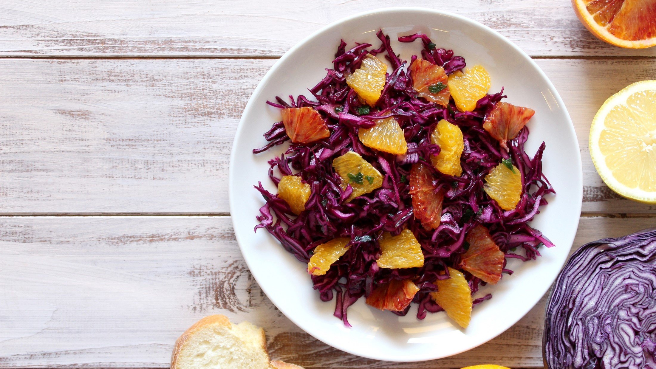 Draufsicht: Ein weißer Teller mit Rotkohlsalat mit Orangen auf einem weißen Untergrund aus Holz. drum herum liegen drei halbierte Zitrusfrüchte.