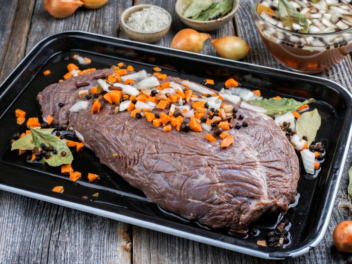 Draufsicht, schräg: Ein Bräter mit einem eigelegten Sauerbraten auf einem Untergrund aus groben Holzdielen. Der Sauerbraten ist in einer Beize, beziehungsweise Marinade. Drum herum liegen frisches Gemüse, Kräuter und Gewürze.