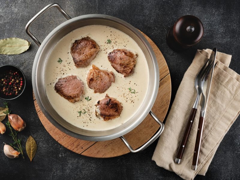 Draufsicht: Eine runde Form aus metall mit zwei henkeln. In der sind Schweinefilet in Apfel-Calvados-Sauce. Drumherum liegen Besteck, ein Tuch sowie frische Zutaten.