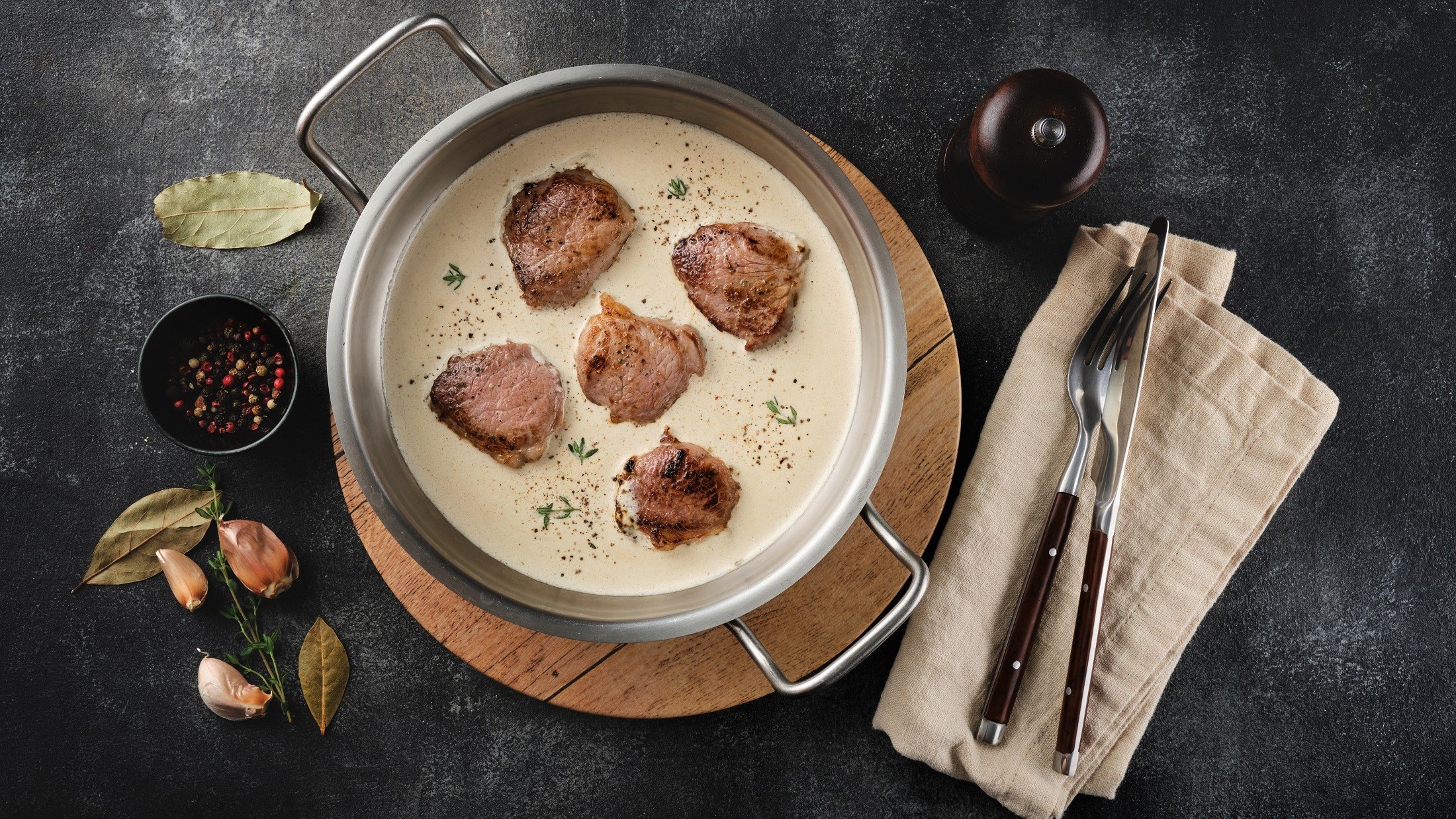 Draufsicht: Eine runde Form aus metall mit zwei henkeln. In der sind Schweinefilet in Apfel-Calvados-Sauce. Drumherum liegen Besteck, ein Tuch sowie frische Zutaten.