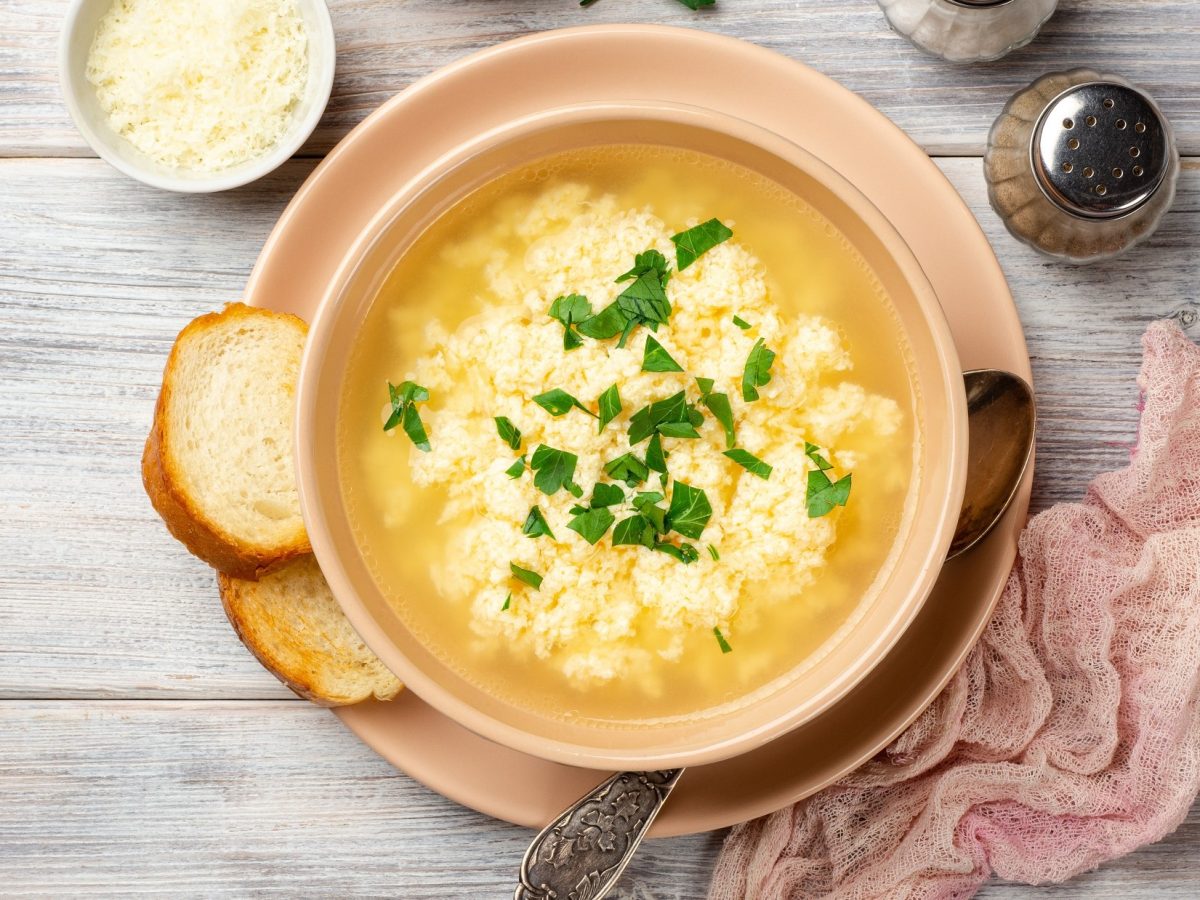 Draufsicht: Eine italienische Stracciatella-Suppe in einer Schale auf einem hellen Untergrund aus Holz. Drumherum liegen eine Serviette und Brotscheiben.