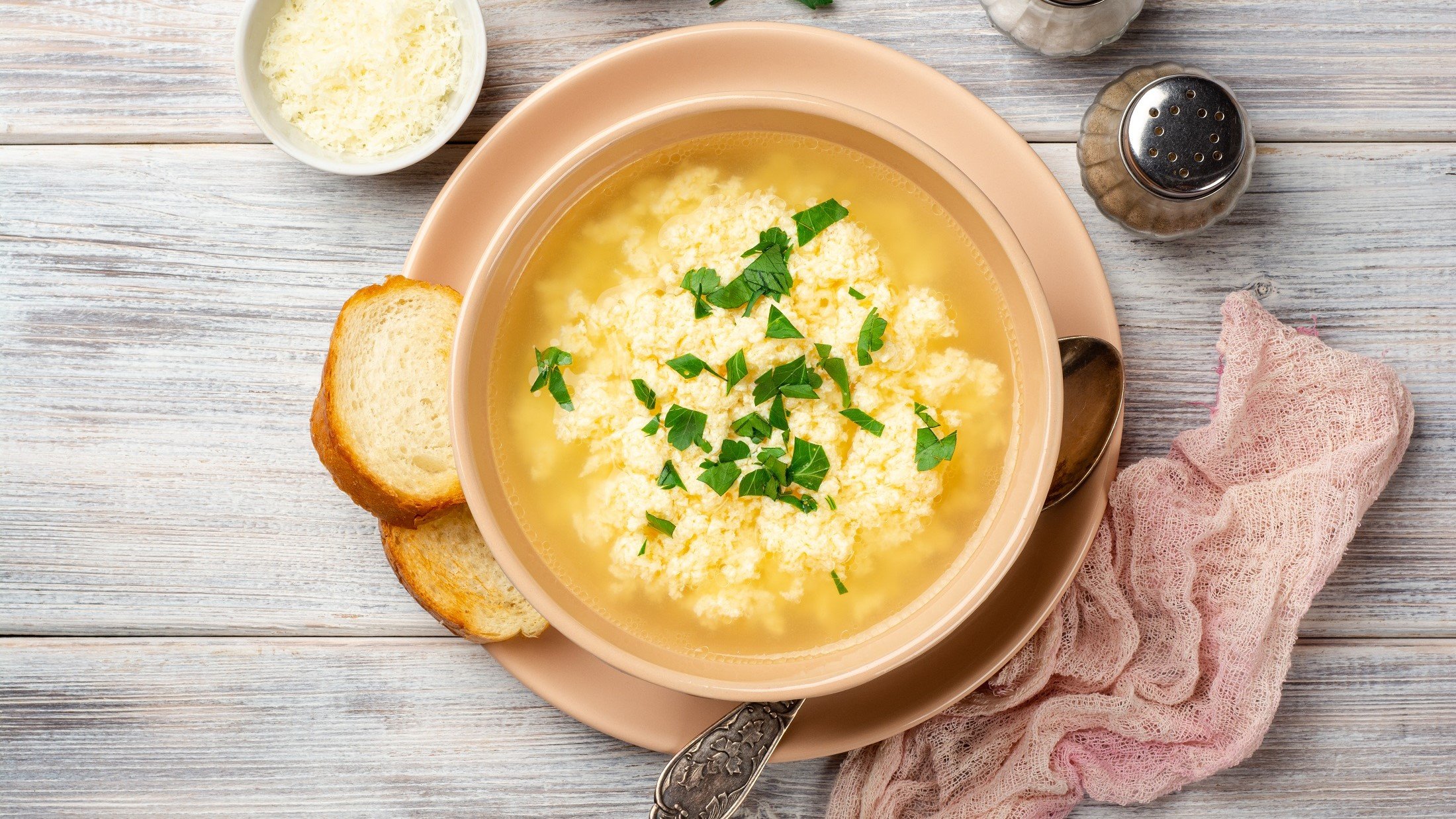 Draufsicht: Eine italienische Stracciatella-Suppe in einer Schale auf einem hellen Untergrund aus Holz. Drumherum liegen eine Serviette und Brotscheiben.