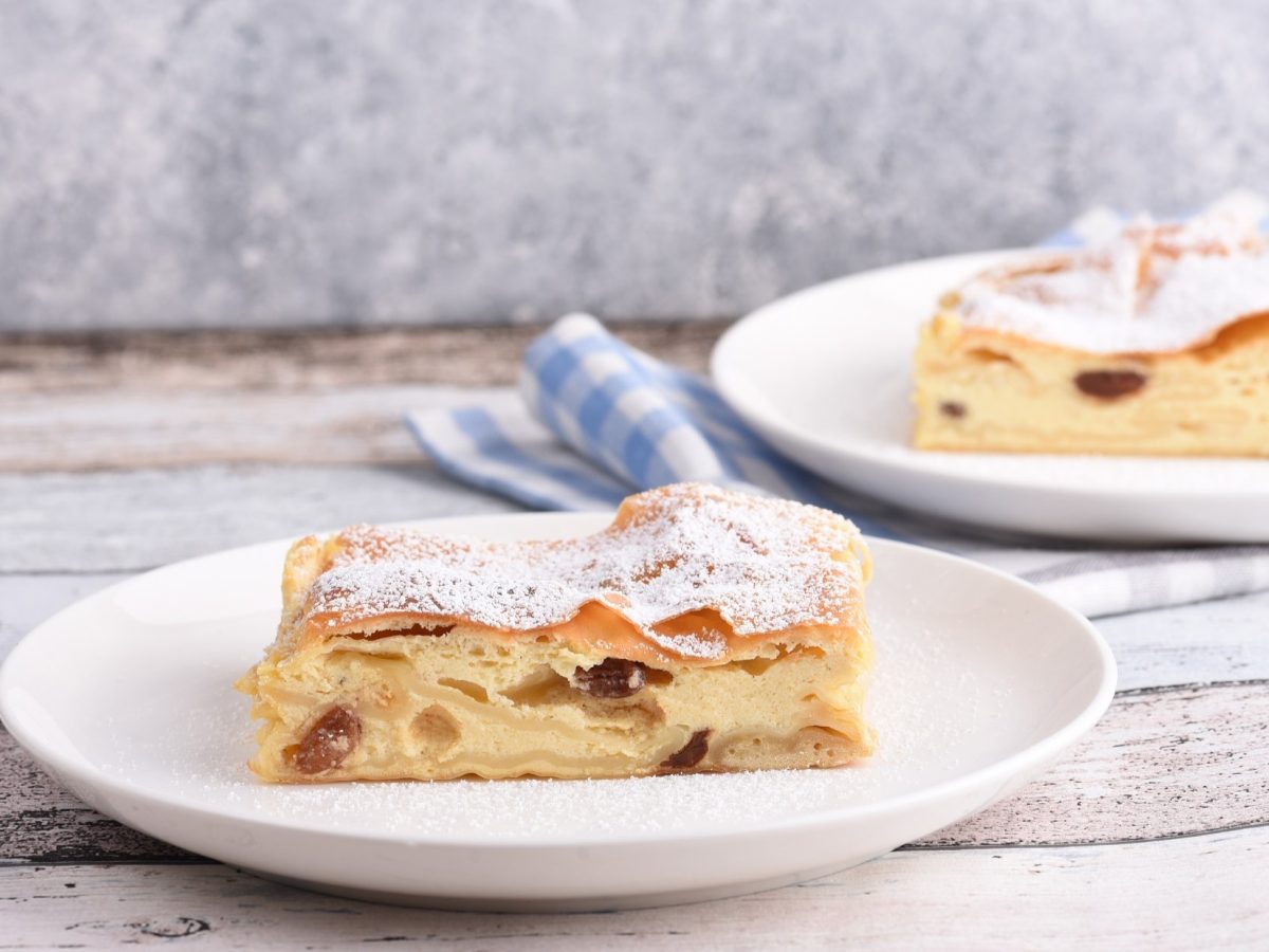 Frontal: Zwei weiße Teller mit Topfenstrudel auf einem hellen Untergrund aus Holz.