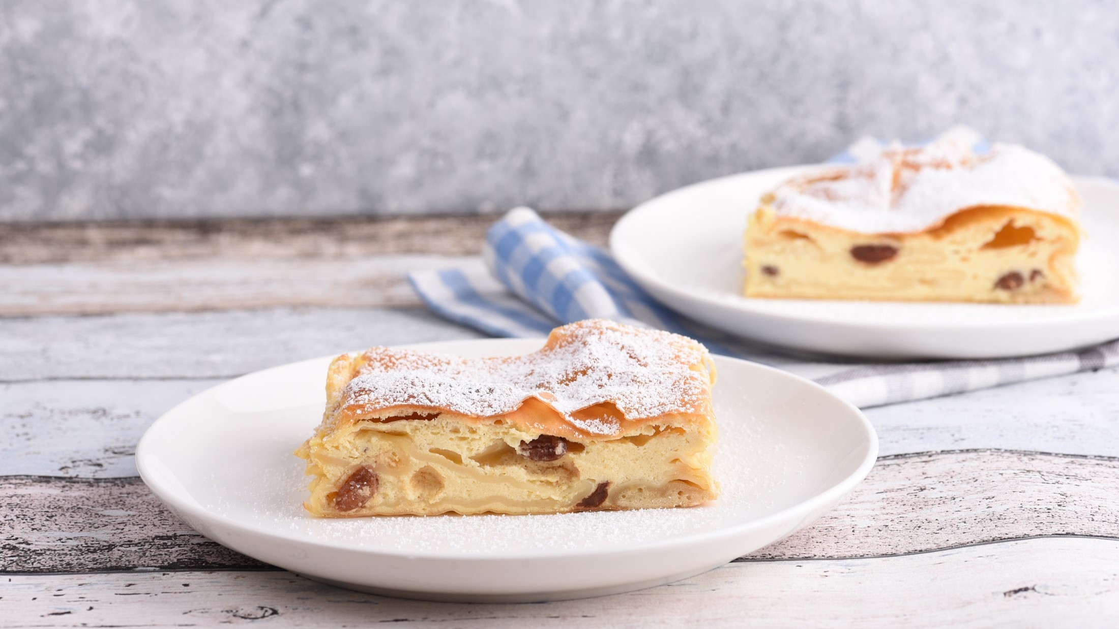 Frontal: Zwei weiße Teller mit Topfenstrudel auf einem hellen Untergrund aus Holz.