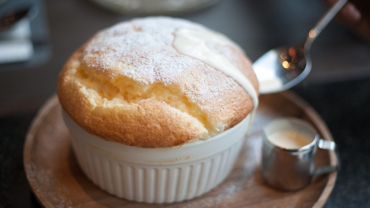 Draufsicht, schräg: Ein selbst gemachtes Vanille-Soufflé auf einem kleinen runden Teller. Daneben liegt ein Löffel und steht ein kleines Kännchen aus Metall mit einer Vanillesauce.