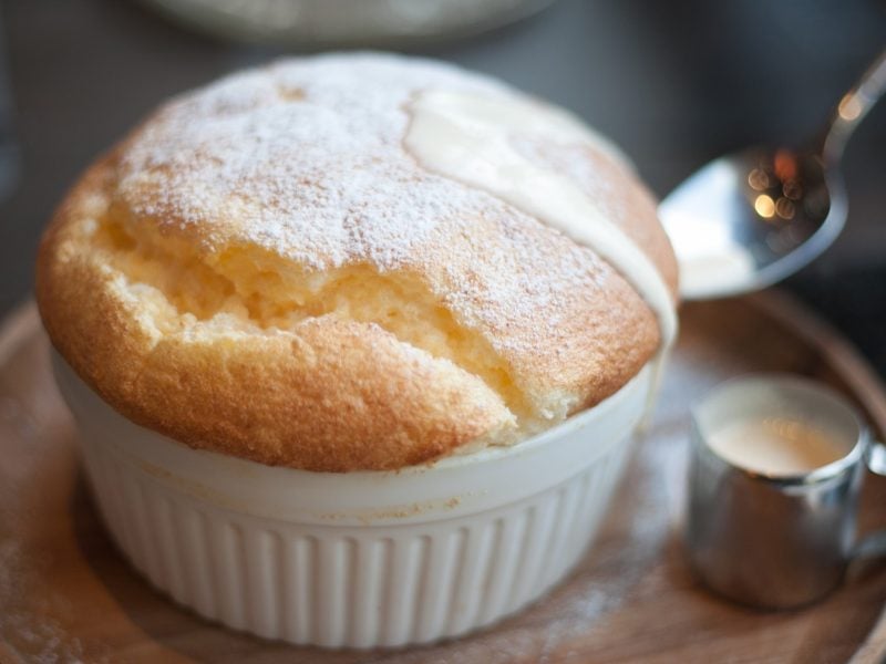 Draufsicht, schräg: Ein selbst gemachtes Vanille-Soufflé auf einem kleinen runden Teller. Daneben liegt ein Löffel und steht ein kleines Kännchen aus Metall mit einer Vanillesauce.