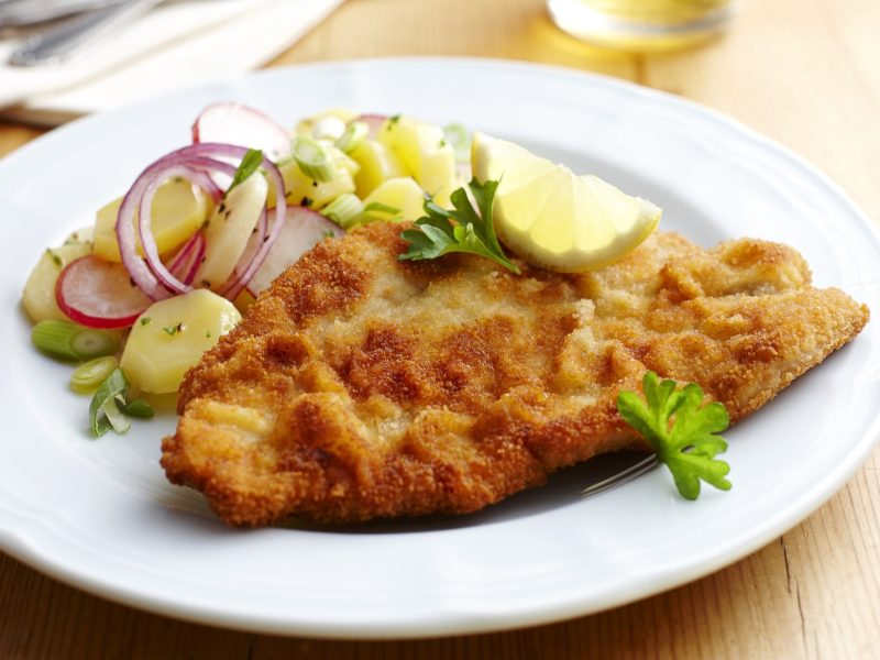 Ein weißer Teller mit einem Wiener Schnitzel und einem Kartoffelsalat auf einem Untergrund aus Holz. Daneben liegt ein Besteck und im Hintergrund ist ein Glas mit einem Getränt.