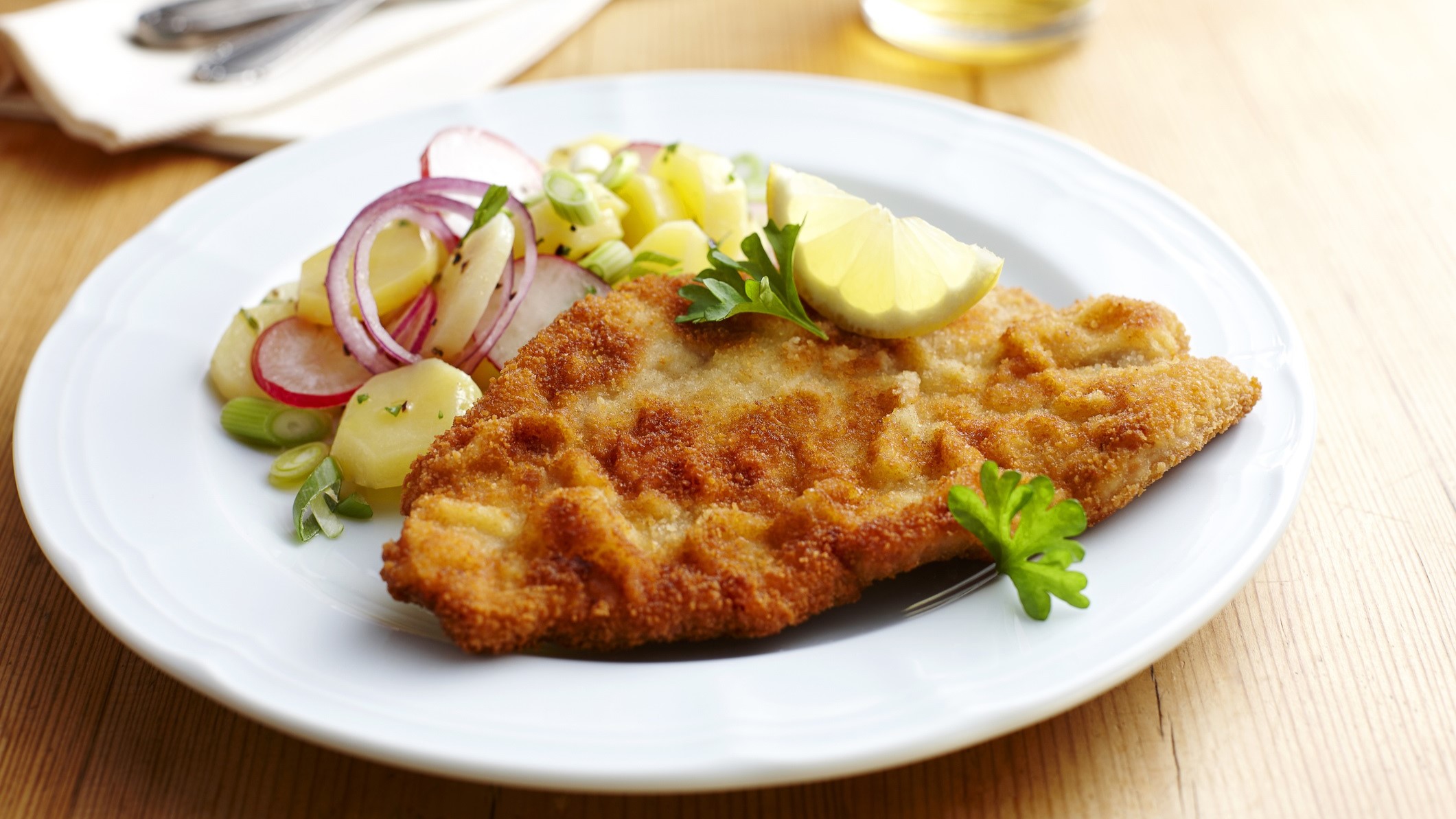 Ein weißer Teller mit einem Wiener Schnitzel und einem Kartoffelsalat auf einem Untergrund aus Holz. Daneben liegt ein Besteck und im Hintergrund ist ein Glas mit einem Getränt.