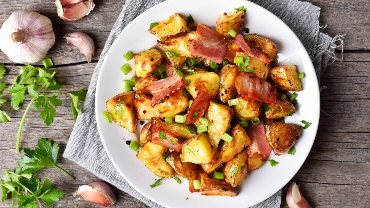 Gebackene Kohlrabistücke mit knusprigem Speck und Frühlingszwiebeln auf einem weißen Teller, umgeben von frischen Kräutern und Knoblauch.