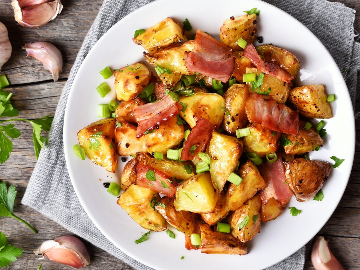 Gebackene Kohlrabistücke mit knusprigem Speck und Frühlingszwiebeln auf einem weißen Teller, umgeben von frischen Kräutern und Knoblauch.