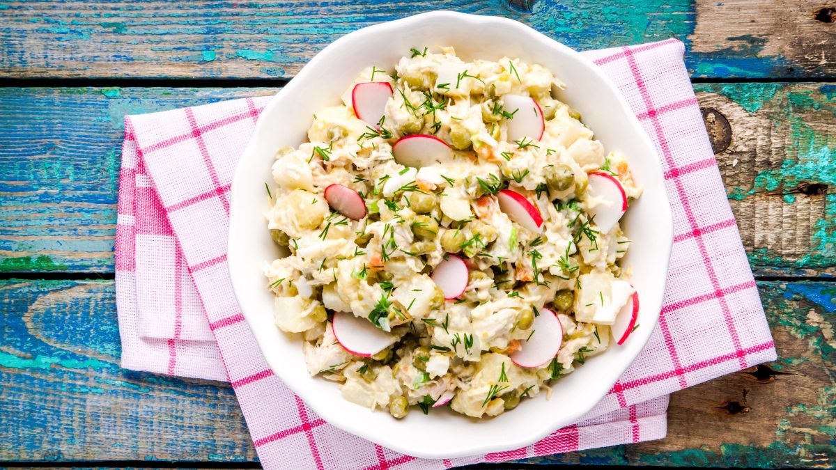 Ein Kohlrabisalat mit Radieschen, Erbsen und Dill in einer weißen Schüssel auf einem bunten Holztisch mit kariertem Tuch.