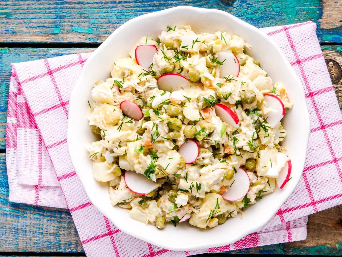 Ein Kohlrabisalat mit Radieschen, Erbsen und Dill in einer weißen Schüssel auf einem bunten Holztisch mit kariertem Tuch.