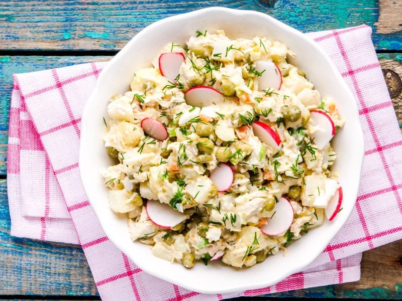 Ein Kohlrabisalat mit Radieschen, Erbsen und Dill in einer weißen Schüssel auf einem bunten Holztisch mit kariertem Tuch.