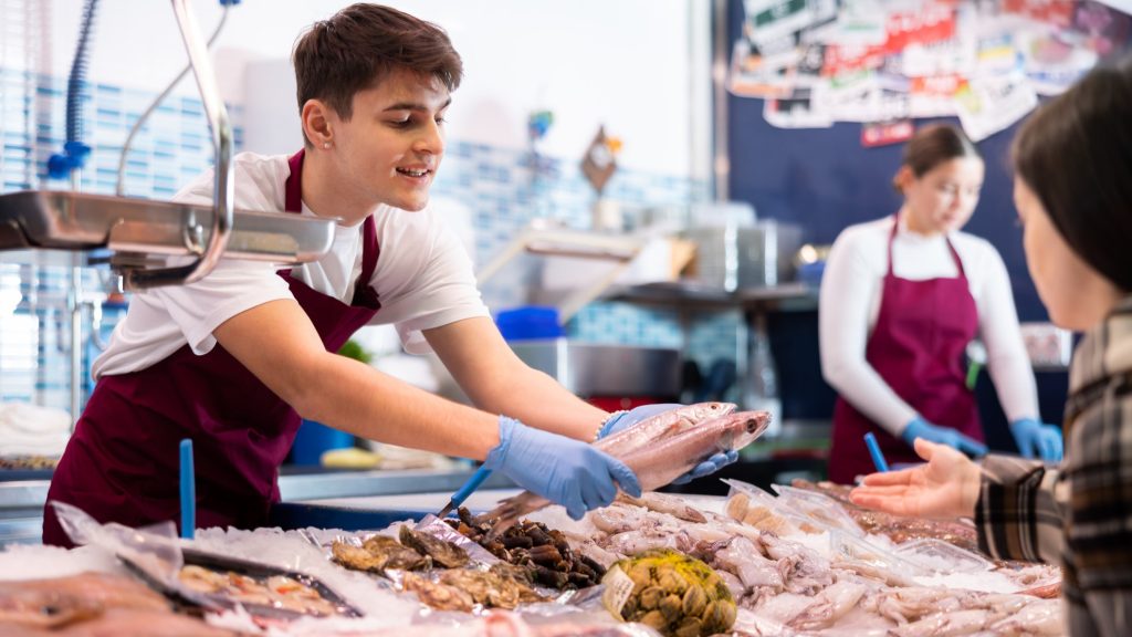 Fischhändler über eine Auslage, der einer Frau Fisch zum Kaufen anbietet.