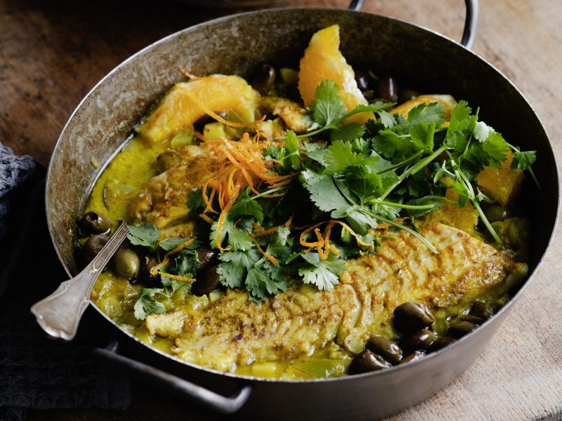 Ein gusseiserner Topf mit der Fisch-Tajine auf beigem Untergrund von schräg oben fotografiert.