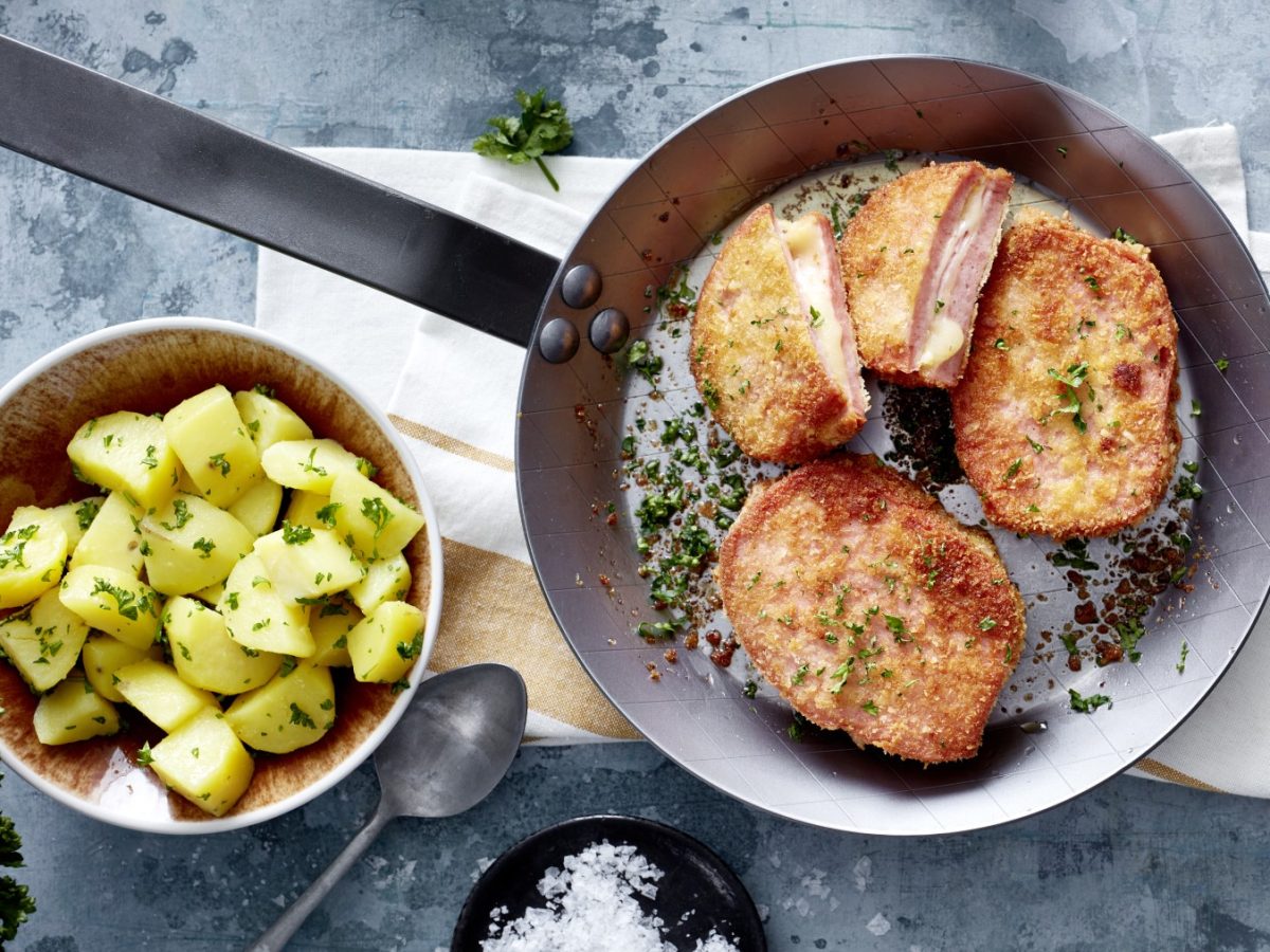 Fleischkäse-Cordon-bleu in einer Pfanne, daneben Kartoffeln in einer Schüssel auf hellem Untergrund.