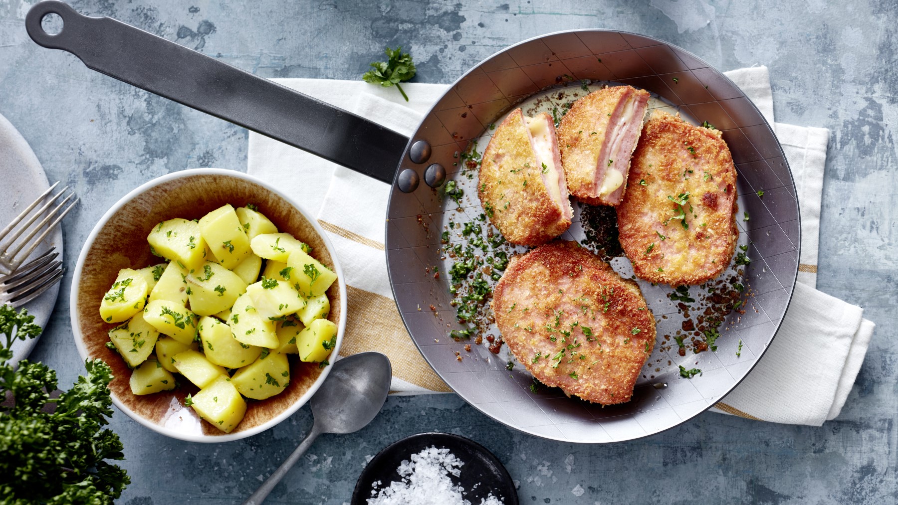 Fleischkäse-Cordon-bleu in einer Pfanne, daneben Kartoffeln in einer Schüssel auf hellem Untergrund.