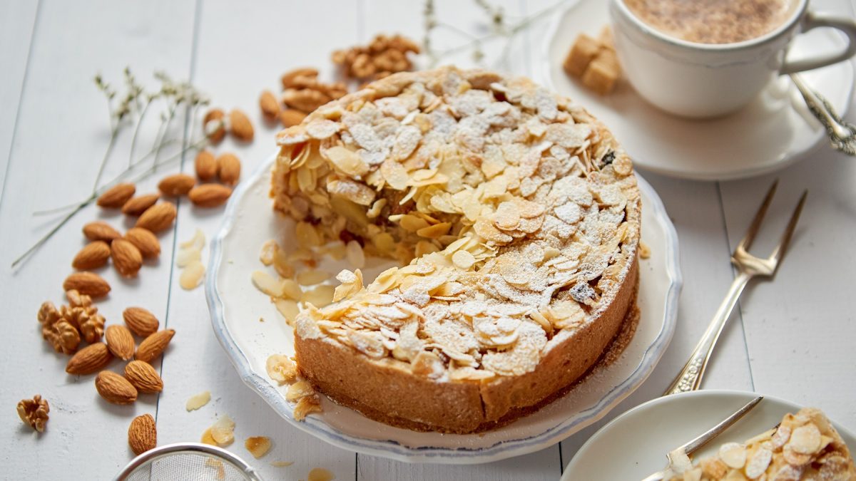 Der Apfelkuchen mit Mandelkruste auf einem weißen Teller und hellem Tisch. Drumherum verteilt sind Mandeln sowie Kaffee und Kuchengeschirr mit einem Stück vom Kuchen.