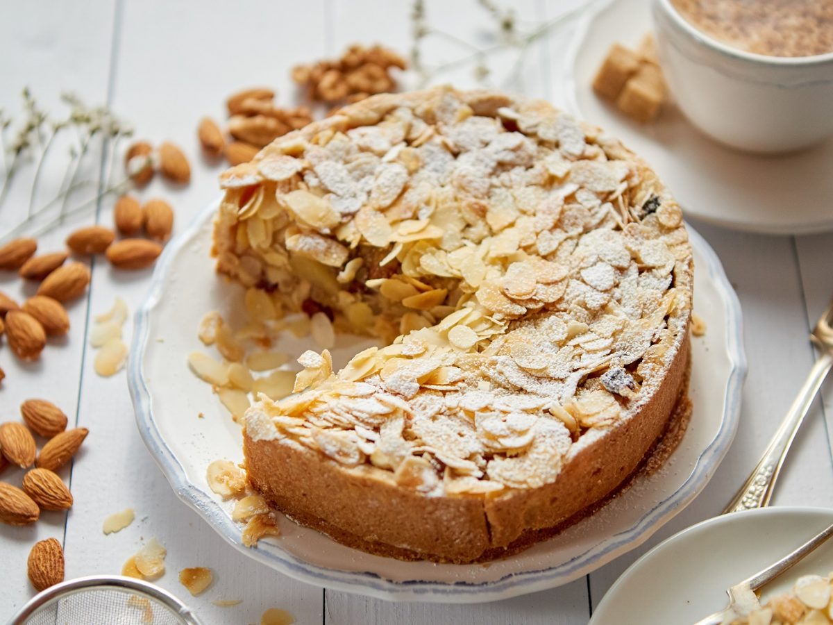 Der Apfelkuchen mit Mandelkruste auf einem weißen Teller und hellem Tisch. Drumherum verteilt sind Mandeln sowie Kaffee und Kuchengeschirr mit einem Stück vom Kuchen.