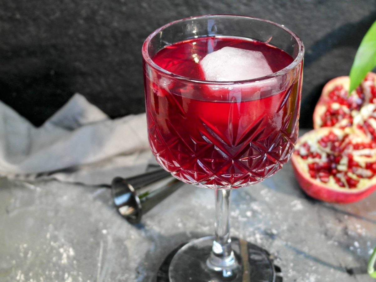 Ein Glas mit dem Granatapfel-Spritz auf einem grauen Steintisch, mit halbiertem Granatapfel, grünen Blättern und schwarzem Hintergrund.