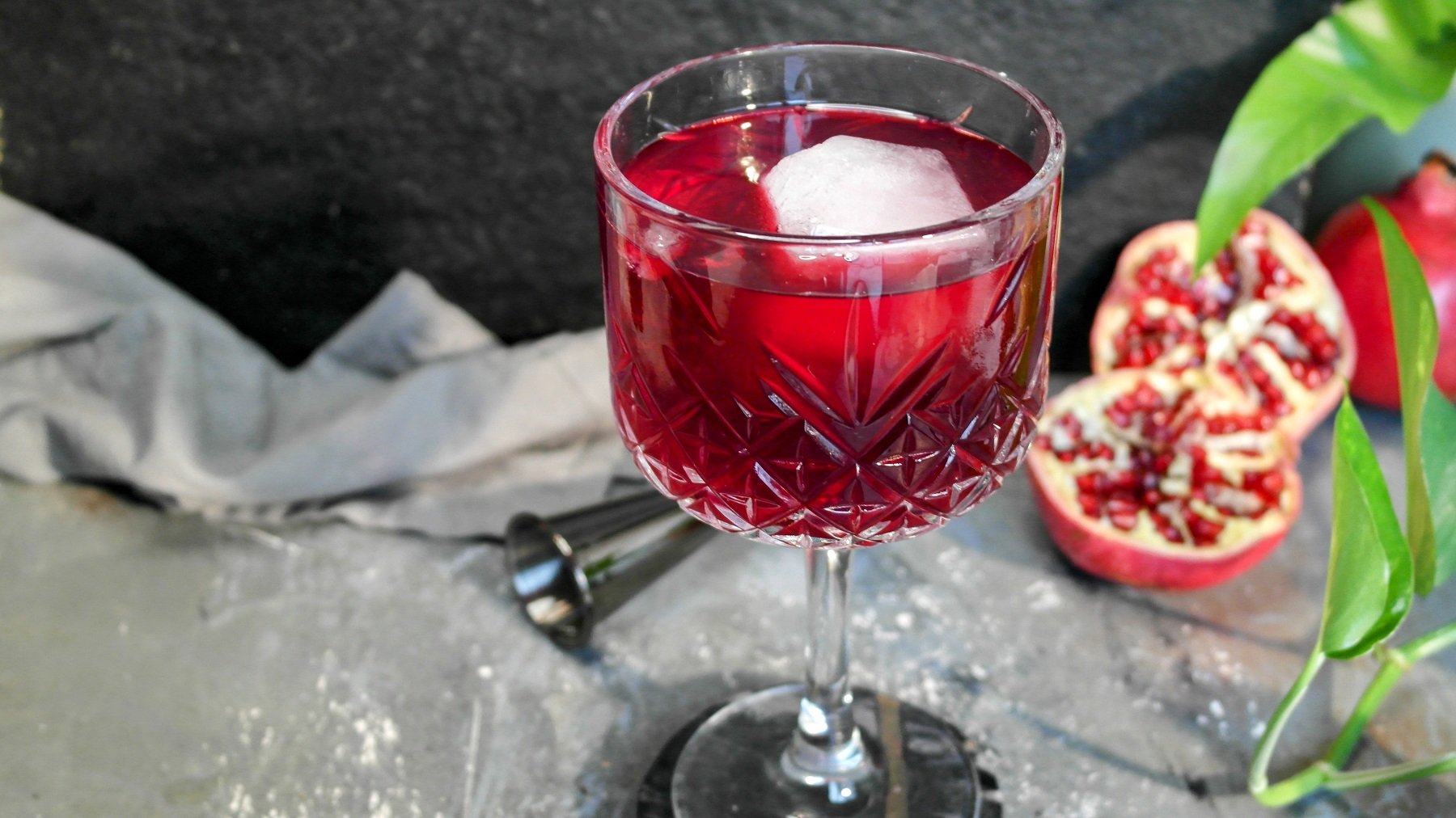 Ein Glas mit dem Granatapfel-Spritz auf einem grauen Steintisch, mit halbiertem Granatapfel, grünen Blättern und schwarzem Hintergrund.