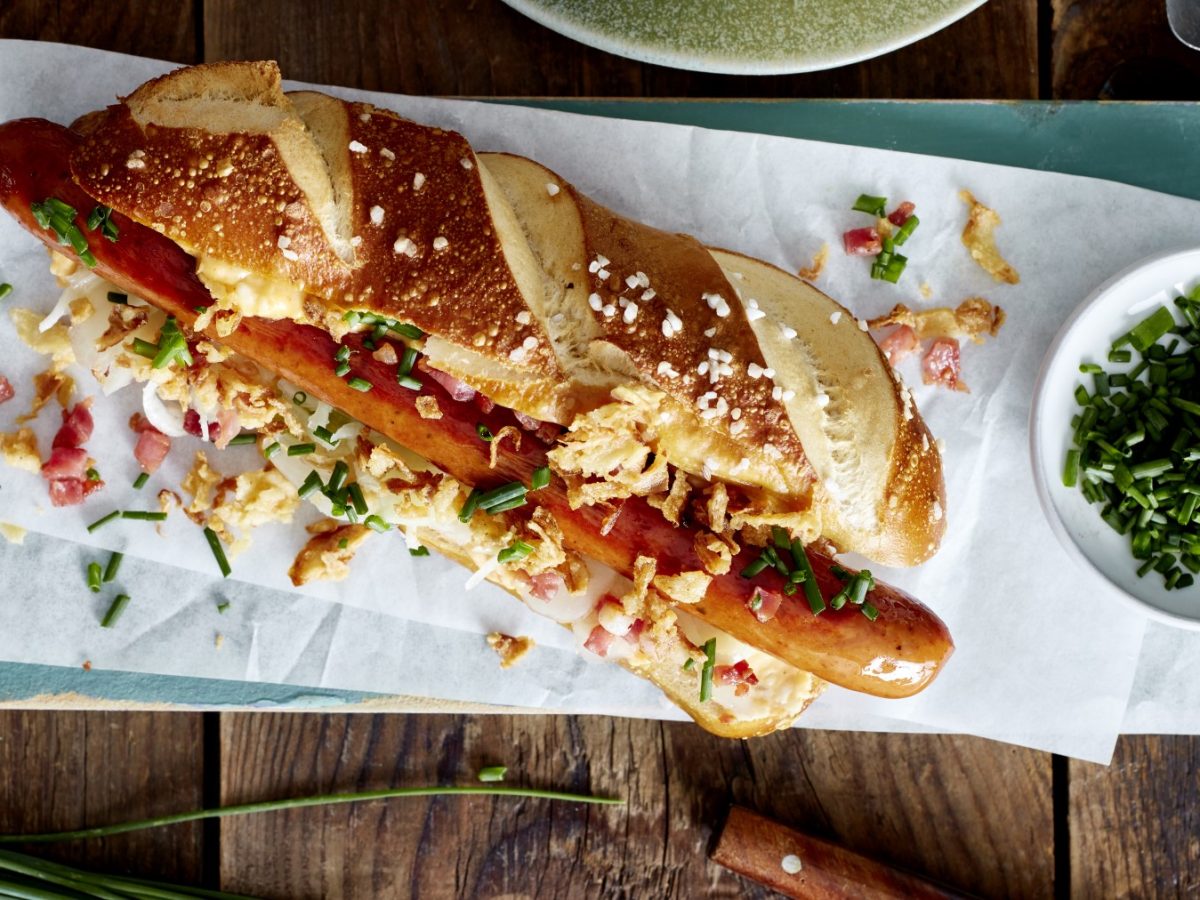 Hotdog auf bayerische Art auf einem hellblauen Brett, rechts davon ein Schüsselchen mit Schnittlauch. Der Hintergrund ist hölzern.