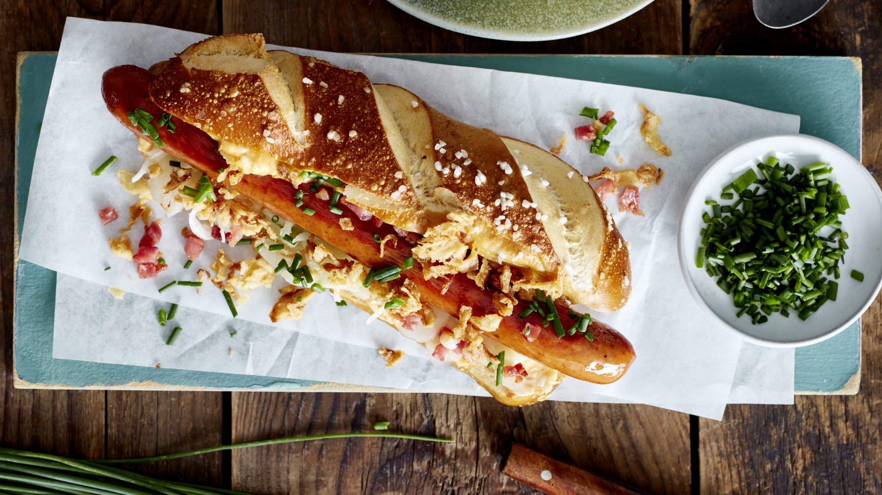 Hotdog auf bayerische Art auf einem hellblauen Brett, rechts davon ein Schüsselchen mit Schnittlauch. Der Hintergrund ist hölzern.