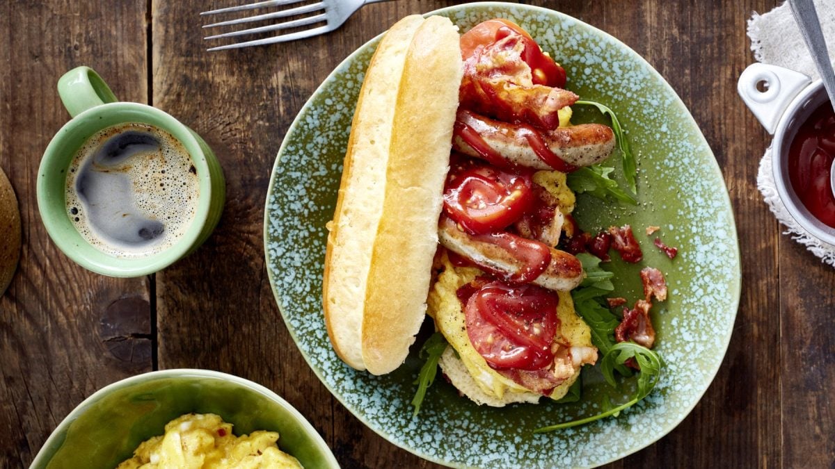 Draufsicht: Hotdog mit Bacon und Rührei auf grünem Teller, daneben Tasse mit Kaffee auf Holzuntergrund.