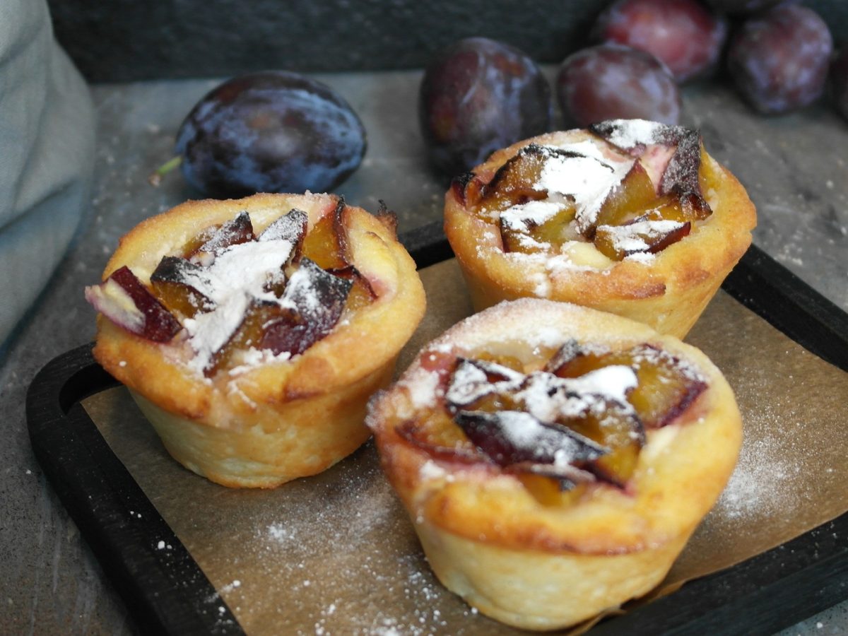 Drei Hüttenkäse-Muffins mit den Zwetschgen auf einem schwarzen Brett mit Zwetschgen und einem grauen Tuch im Hintergrund.