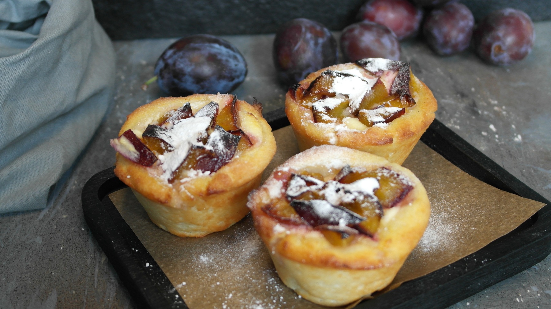 Drei Hüttenkäse-Muffins mit den Zwetschgen auf einem schwarzen Brett mit Zwetschgen und einem grauen Tuch im Hintergrund.