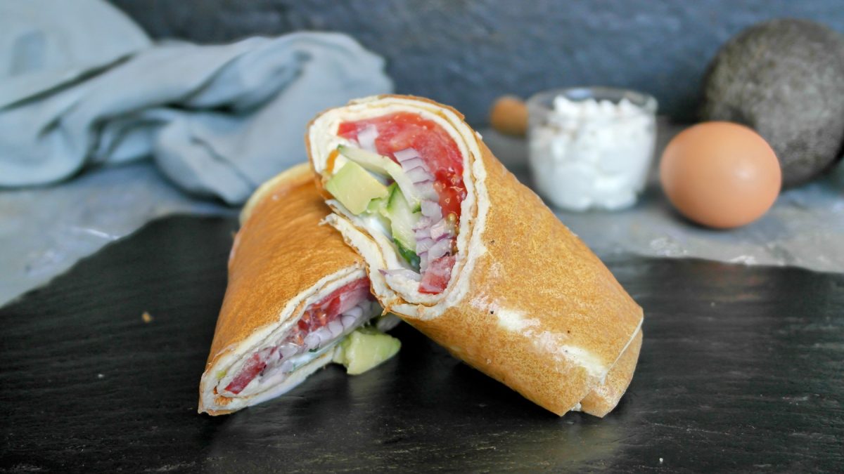 Der angeschnittene Wrap auf einer schwarzen Schiefertafel mit grauem Hintergrund. Außerdem Hüttenkäse, Ei und Avocado unscharf im Hintergrund erkennbar.