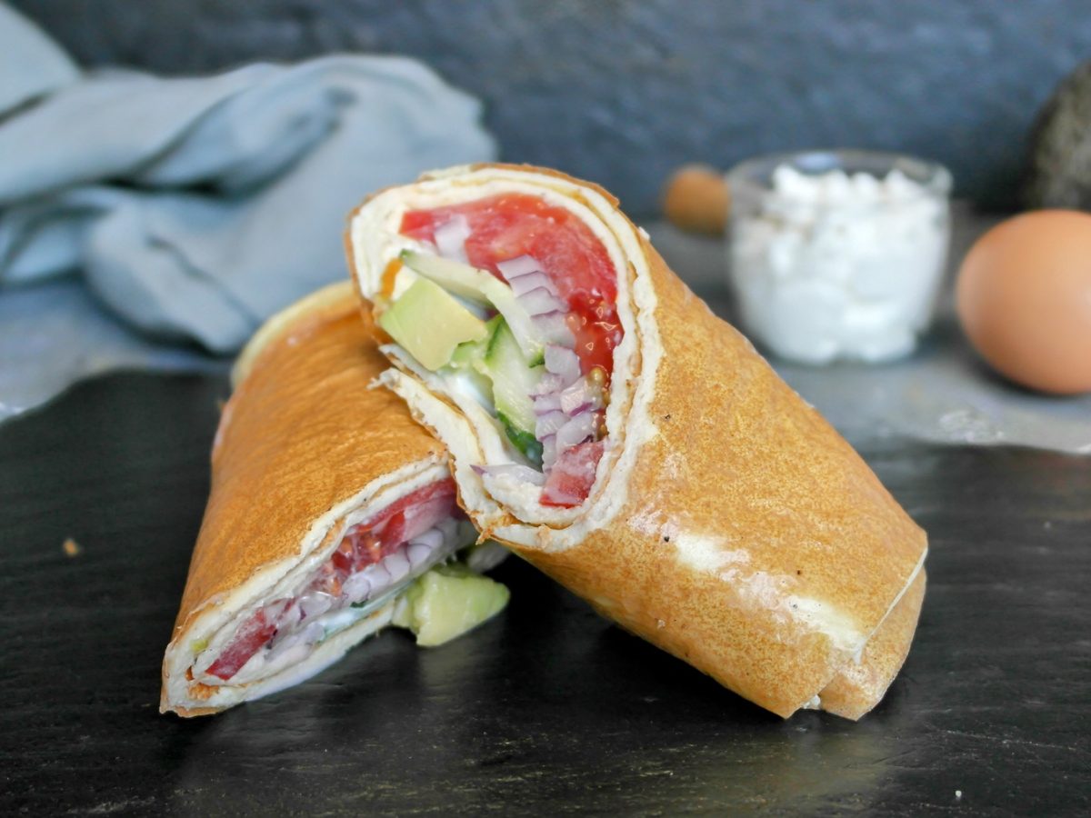 Der angeschnittene Wrap auf einer schwarzen Schiefertafel mit grauem Hintergrund. Außerdem Hüttenkäse, Ei und Avocado unscharf im Hintergrund erkennbar.