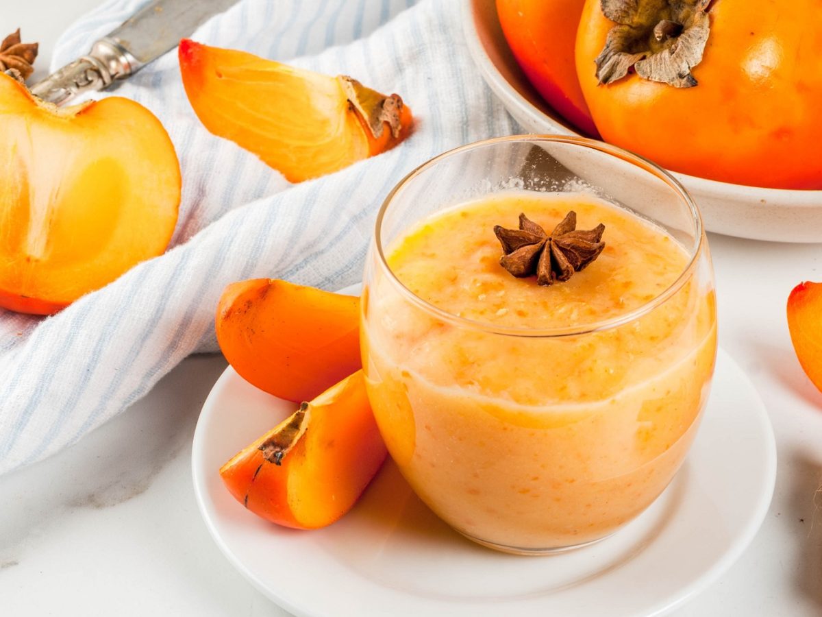 Ein Glas mit dem Kaki-Smoothie und im Hintergrund angeshcnittene und ganze Kaki. Alles auf einem hellen Tisch mit weißem Tuch von schräg oben fotografiert.