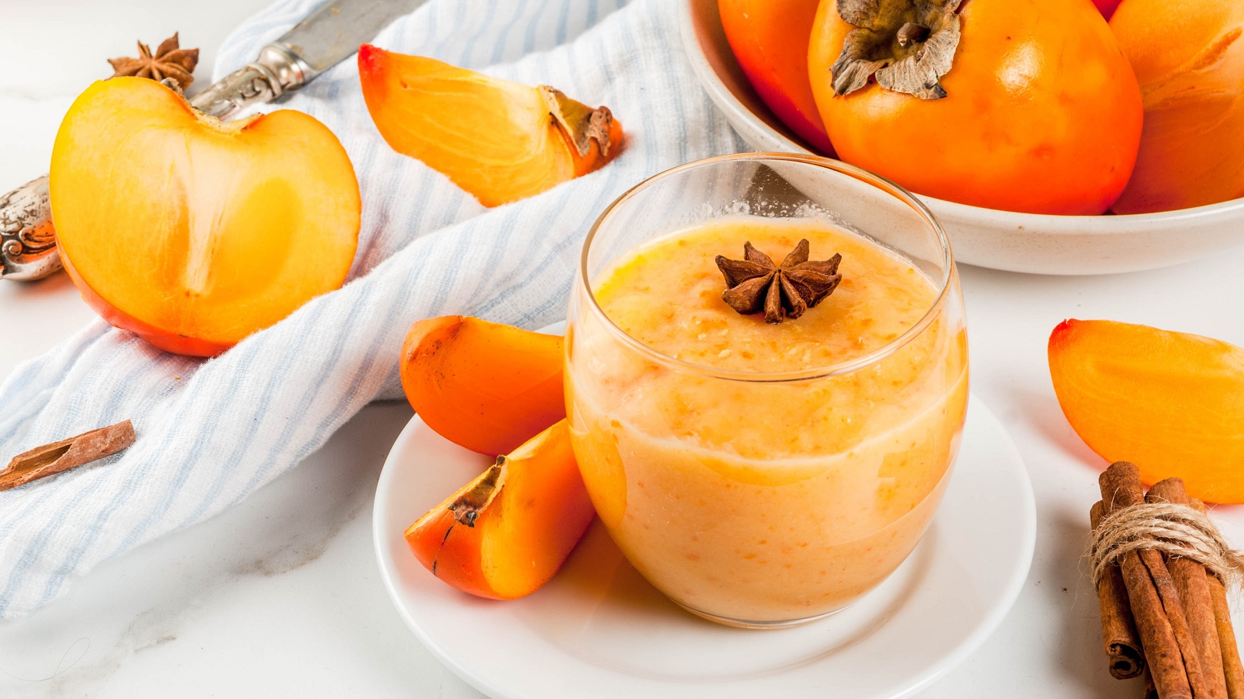 Ein Glas mit dem Kaki-Smoothie und im Hintergrund angeshcnittene und ganze Kaki. Alles auf einem hellen Tisch mit weißem Tuch von schräg oben fotografiert.