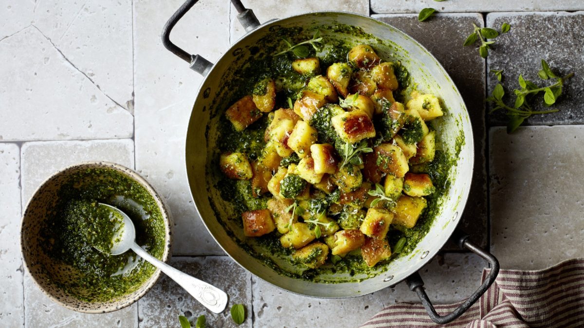 Knusper-Gnocchetti in Rucola-Kürbiskern-Pesto in einer Pfanne, daneben eine Schüssel mit Pesto und Löffel auf grauem Stein-Untergrund.