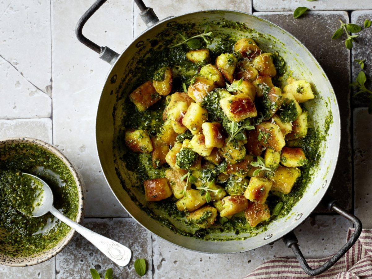 Knusper-Gnocchetti in Rucola-Kürbiskern-Pesto in einer Pfanne, daneben eine Schüssel mit Pesto und Löffel auf grauem Stein-Untergrund.