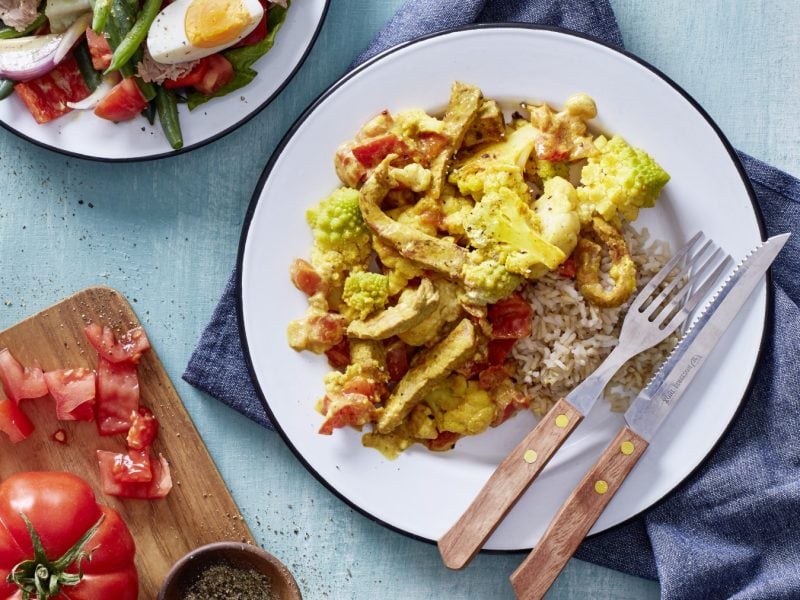 Kohl mit Schnitzel und Curry in einer weißen Schüssel auf hellblauem Hintergrund. Links unten auf dem Bild ein Schneidebrett mit Tomaten und links oben ein Teller mit Salat.