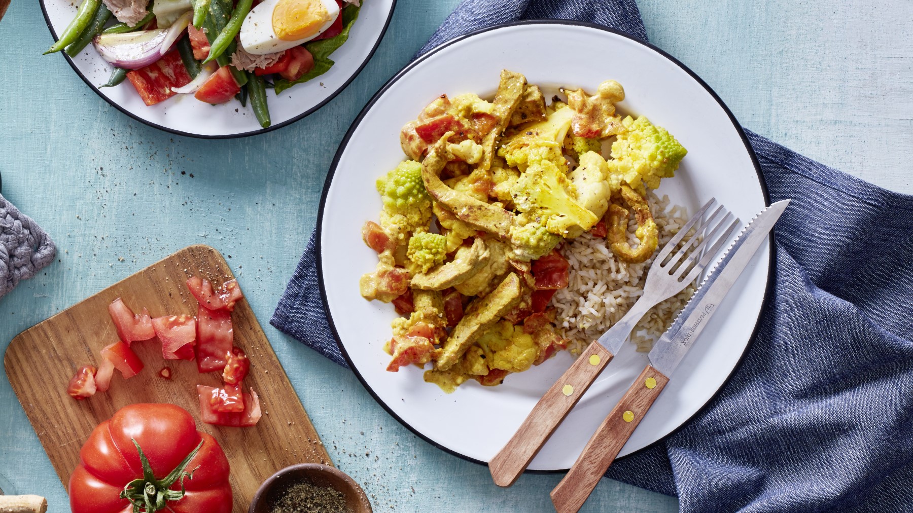 Kohl mit Schnitzel und Curry in einer weißen Schüssel auf hellblauem Hintergrund. Links unten auf dem Bild ein Schneidebrett mit Tomaten und links oben ein Teller mit Salat.