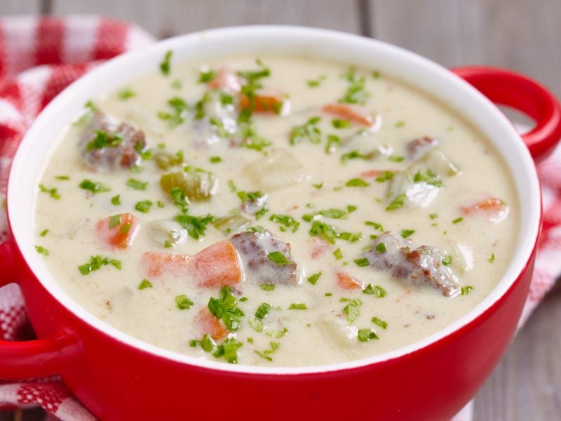 Eine cremige Suppe mit Hackfleisch, Karotten, Sellerie und Petersilie wird in einer roten Schale serviert, umgeben von kariertem Tuch.