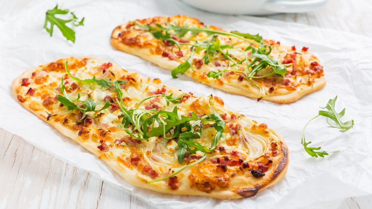 Zwei Flammkuchen auf einem weißen Tisch, mit Rucola bestreut von der Seite fotografiert.